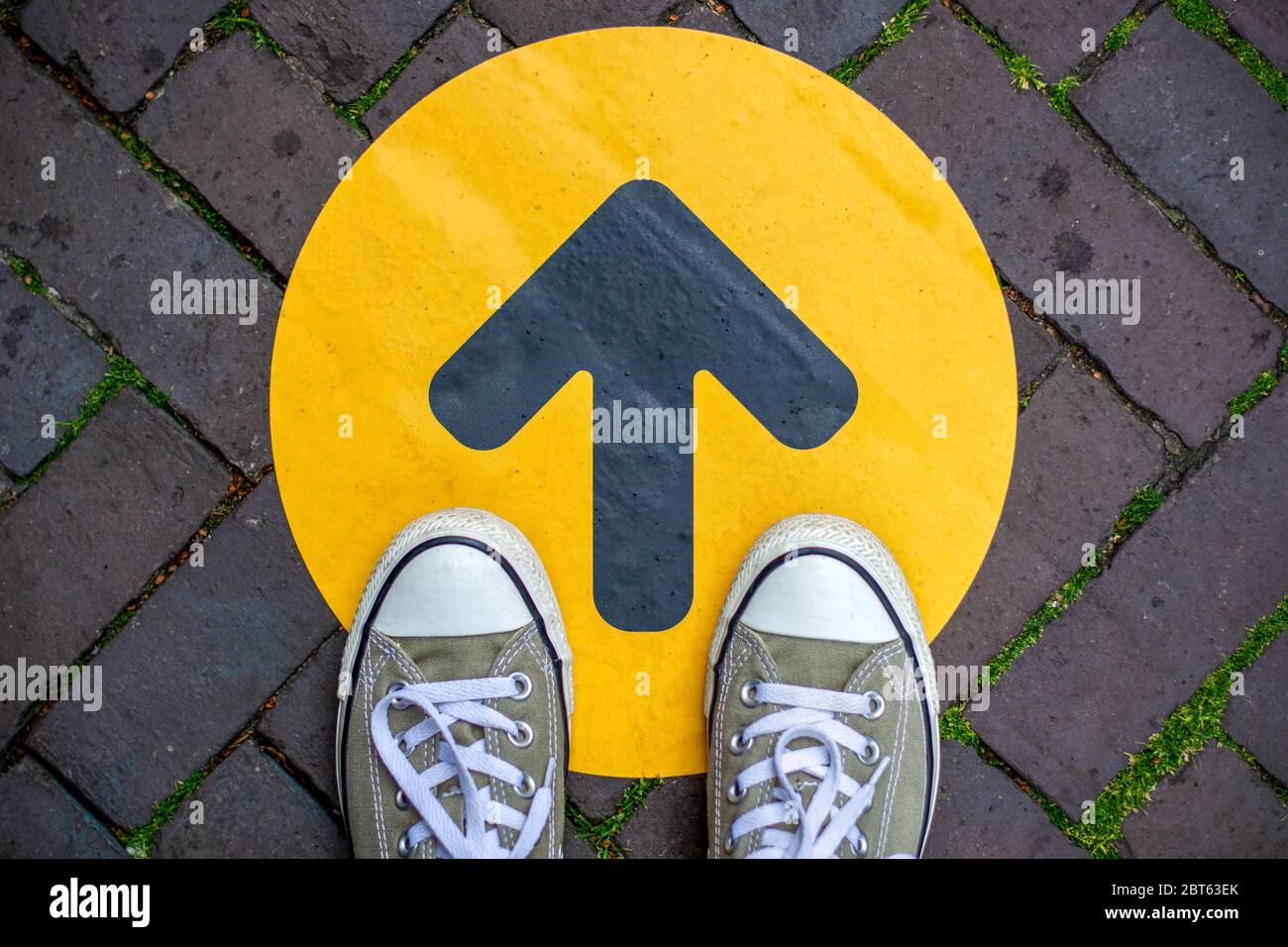 Piedi che indossano sneakers davanti alla freccia sulla strada all'aperto. Freccia di direzione davanti a un negozio per la distanza sociale durante la pandemia di covid-19. Foto Stock