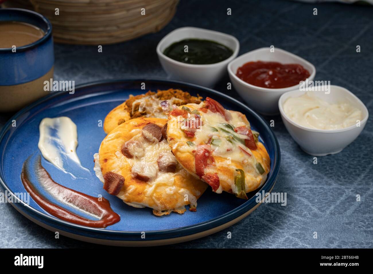 Pizza assortita con salsa di pomodoro, chutney verde e maionese come contorno servito su un piatto blu e una tazza di tè caldo Foto Stock