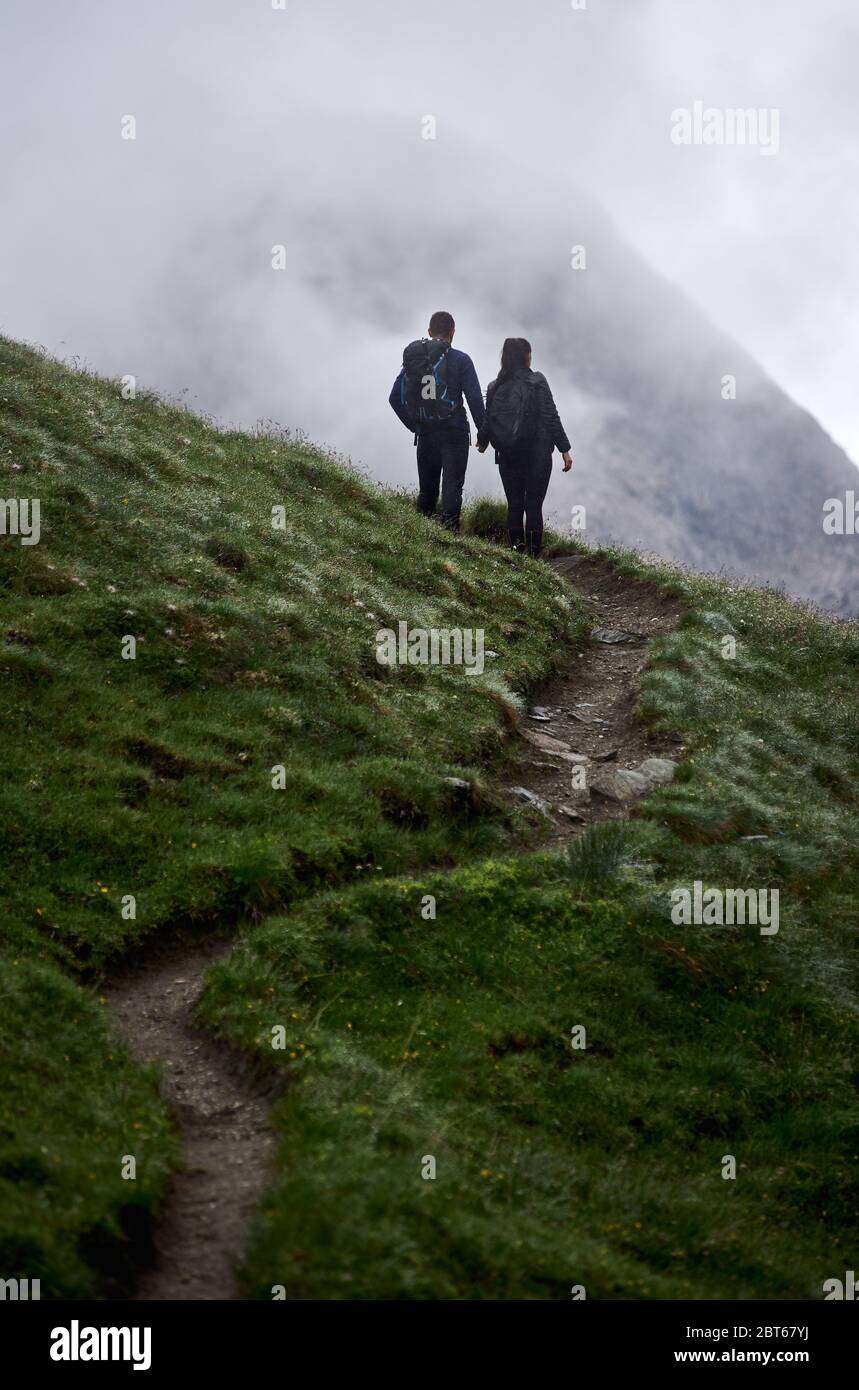 Vista posteriore del viaggiatore maschile e della ragazza che si erge su un percorso erboso collinare e tiene le mani. Coppia amorosa che ammira la vista della montagna nebbiosa mentre si viaggia insieme. Concetto di escursioni e amore. Foto Stock