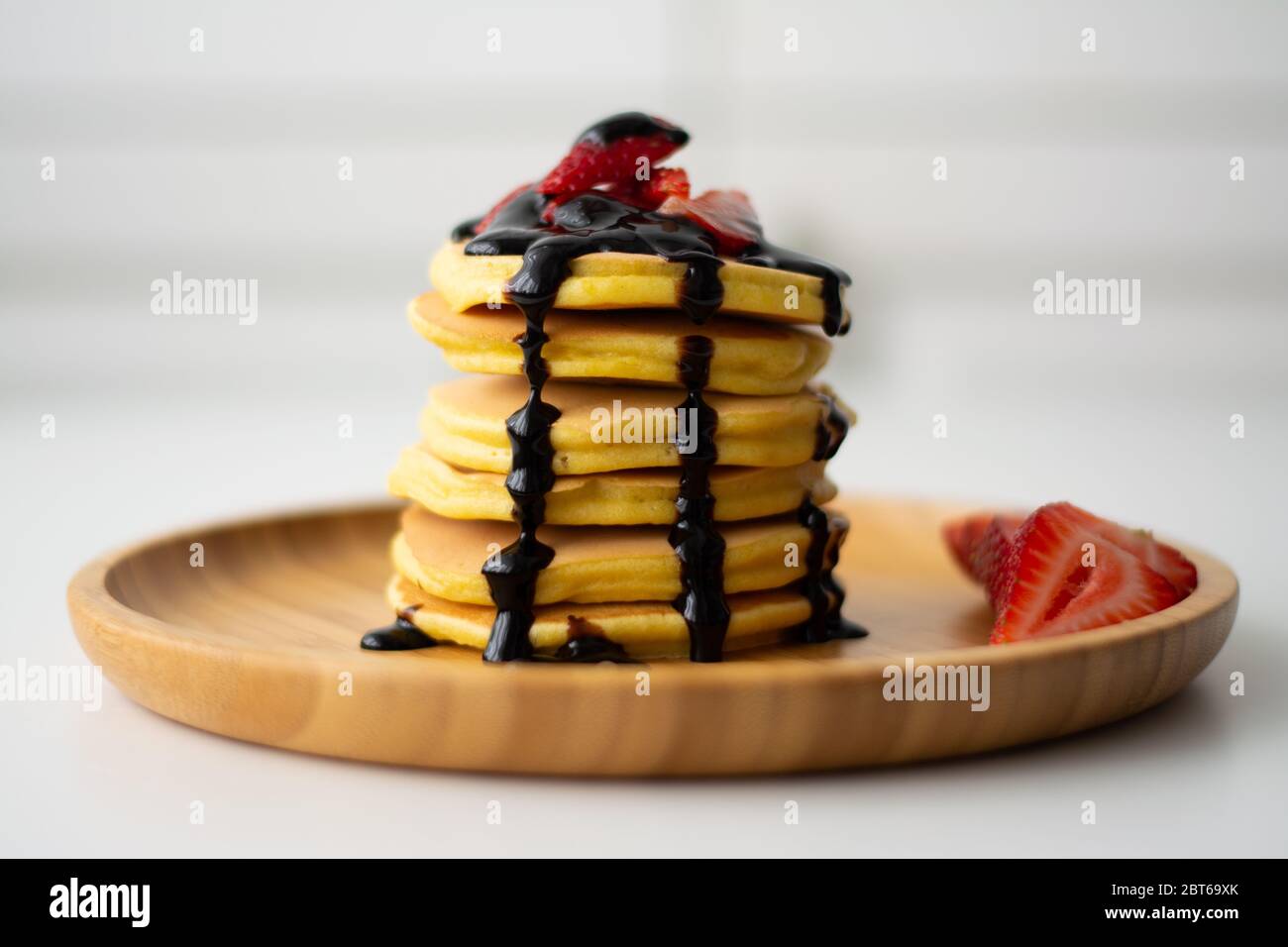 Una pila di frittelle americane soffici per la colazione con fragole e sciroppo di salsa al cioccolato che si versa dall'alto su un piatto di legno con fragola tagliata a fette Foto Stock