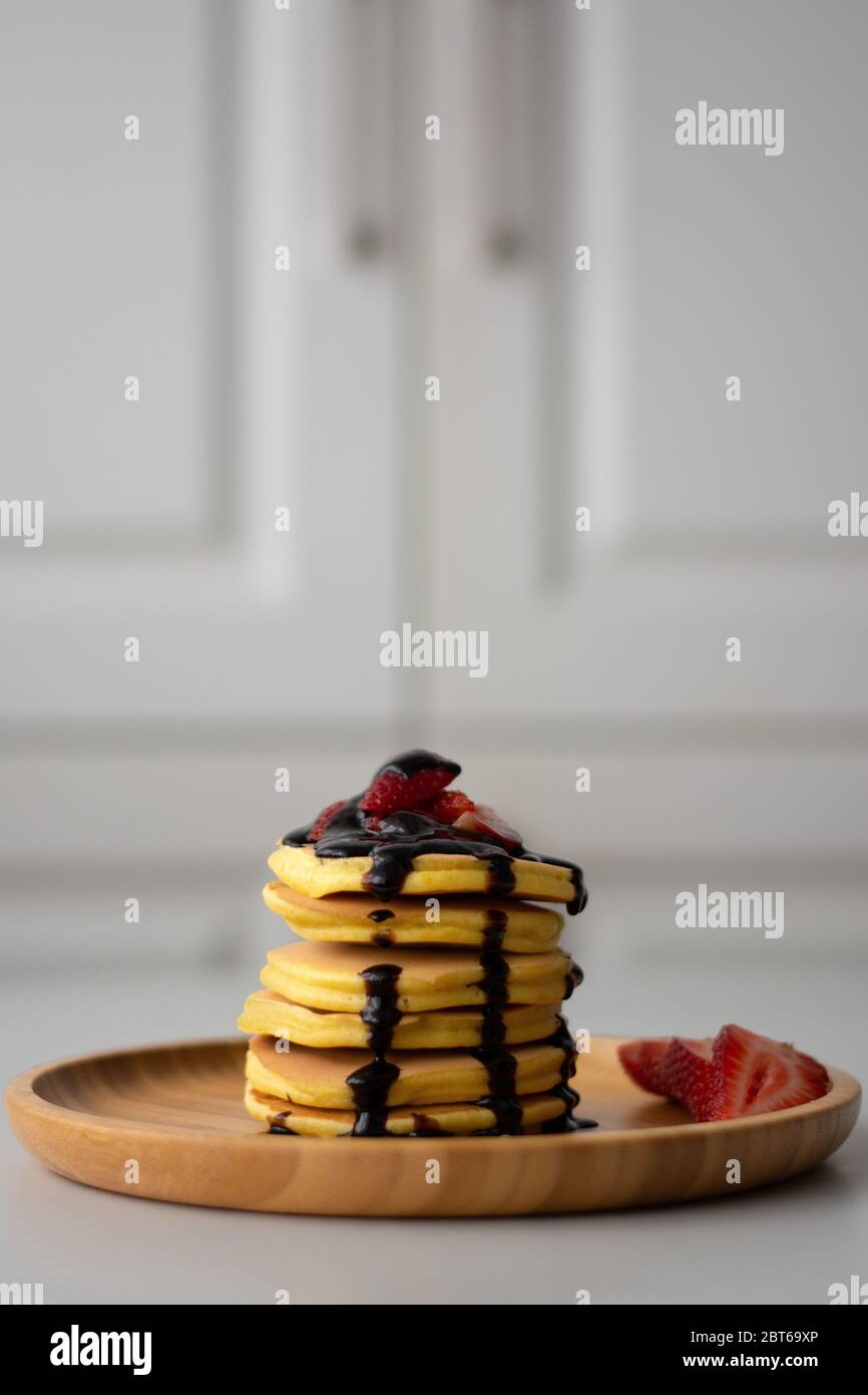 Una pila di frittelle americane soffici per la colazione con fragole e sciroppo di salsa al cioccolato che si versa dall'alto su un piatto di legno con fragola tagliata a fette Foto Stock