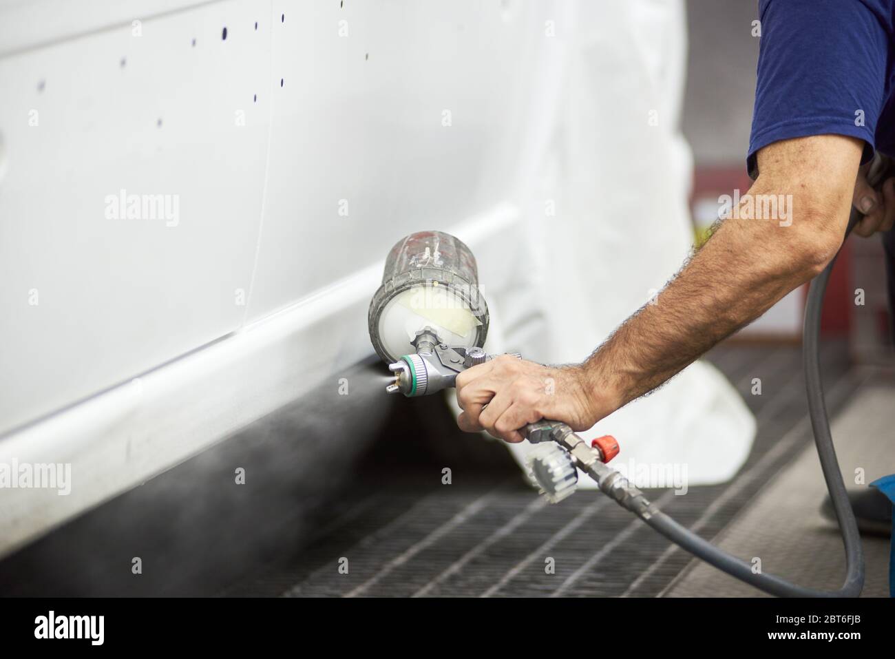 Primo piano della pistola a spruzzo con impugnatura maschio e verniciatura del veicolo in bianco. Gentleman che dimostra la tecnica di verniciatura a spruzzo su automobile. Concetto di servizio automatico e di messa a punto. Foto Stock
