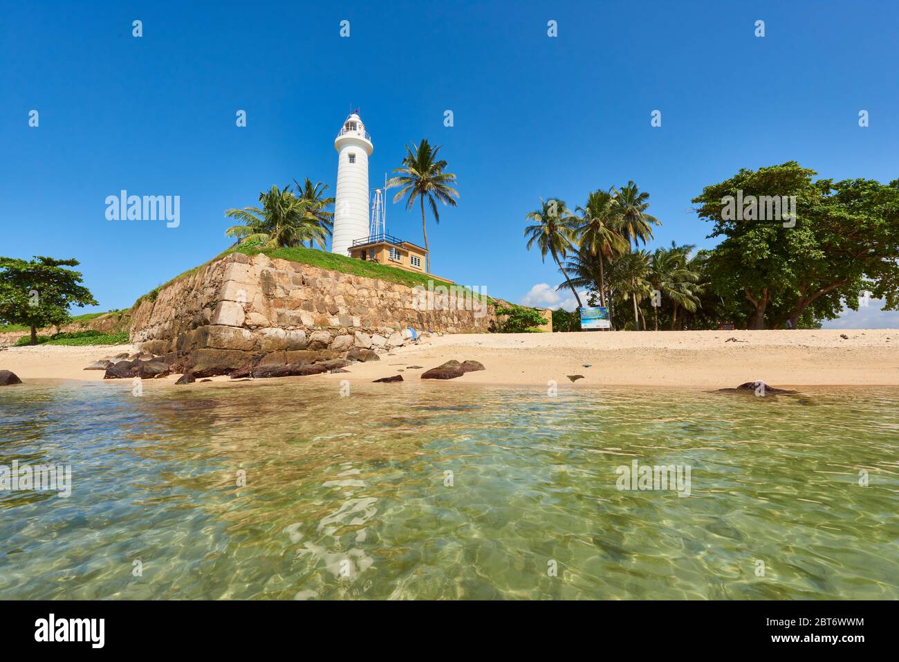 Foto diurna del famoso Faro di Galle circondato da palme da cocco, in piedi alto vicino alle acque cristalline del mare sulla costa. A Fort Galle, Sri L. Foto Stock