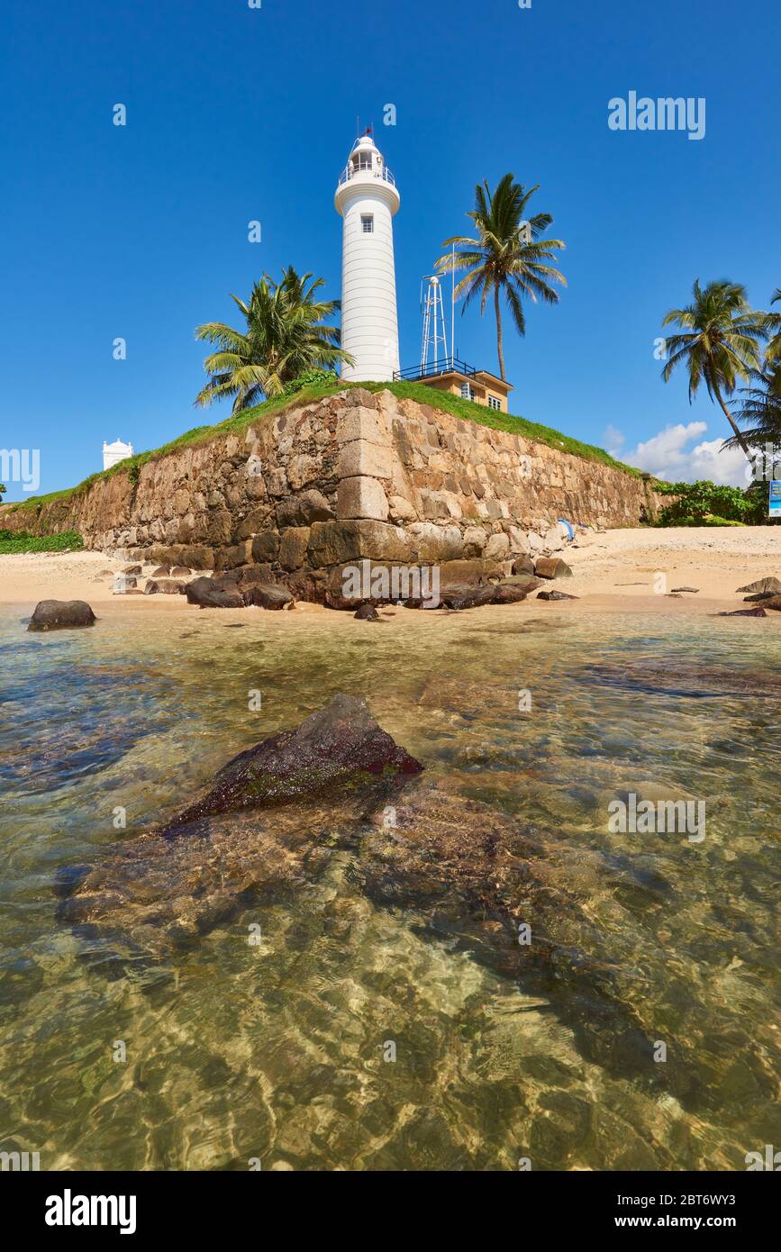 Foto diurna del famoso Faro di Galle circondato da palme da cocco, in piedi alto vicino alle acque cristalline del mare sulla costa. A Fort Galle, Sri L. Foto Stock