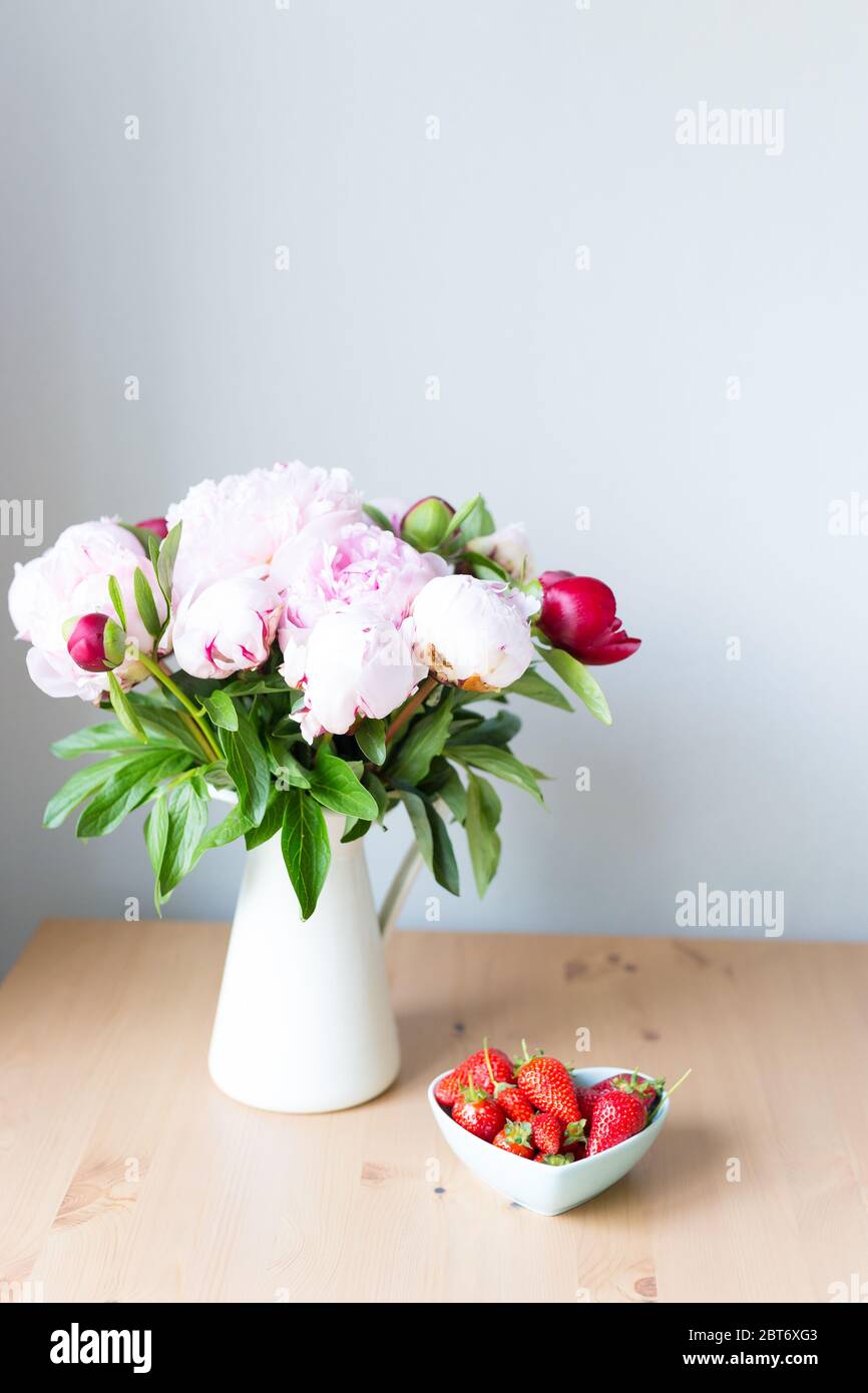 Mazzo di peonie rosa in vaso e fragola sul tavolo di legno . Fiori su un tavolo di legno beige vicino alla finestra. Casa interno. Bellissima Foto Stock