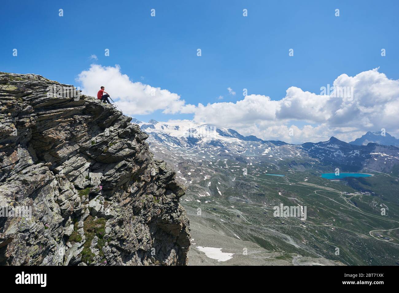 Splendida vista del viaggiatore seduto sul bordo di alta scogliera rocciosa sotto il cielo nuvoloso. Uomo turistico ammirando la vista della valle di montagna con colline e lago blu. Concetto di viaggio, escursioni e alpinismo. Foto Stock