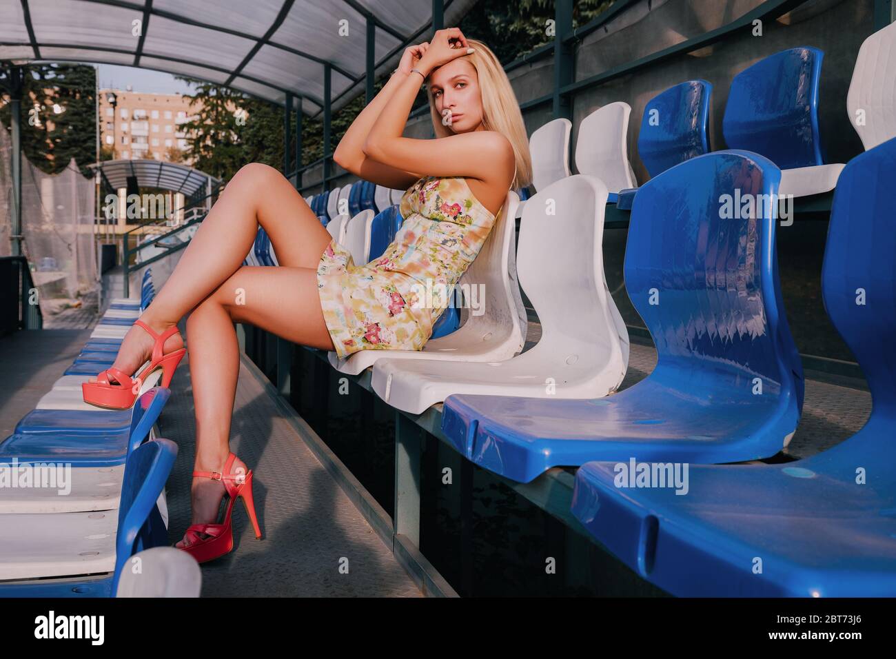 Bella ragazza bionda è seduta su un supporto (tribuna). Indossa un vestito corto leggero e tacchi alti rossi. È soleggiata. Foto Stock