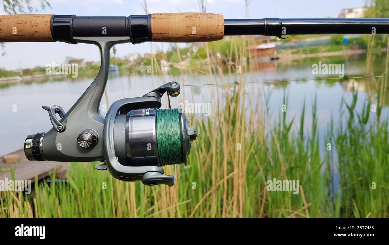 Mulinello da pesca su un comel rotante primo piano sullo sfondo di un lago o di un fiume. Bobina con lenza verde su sfondo naturale. Equi del pescatore Foto Stock