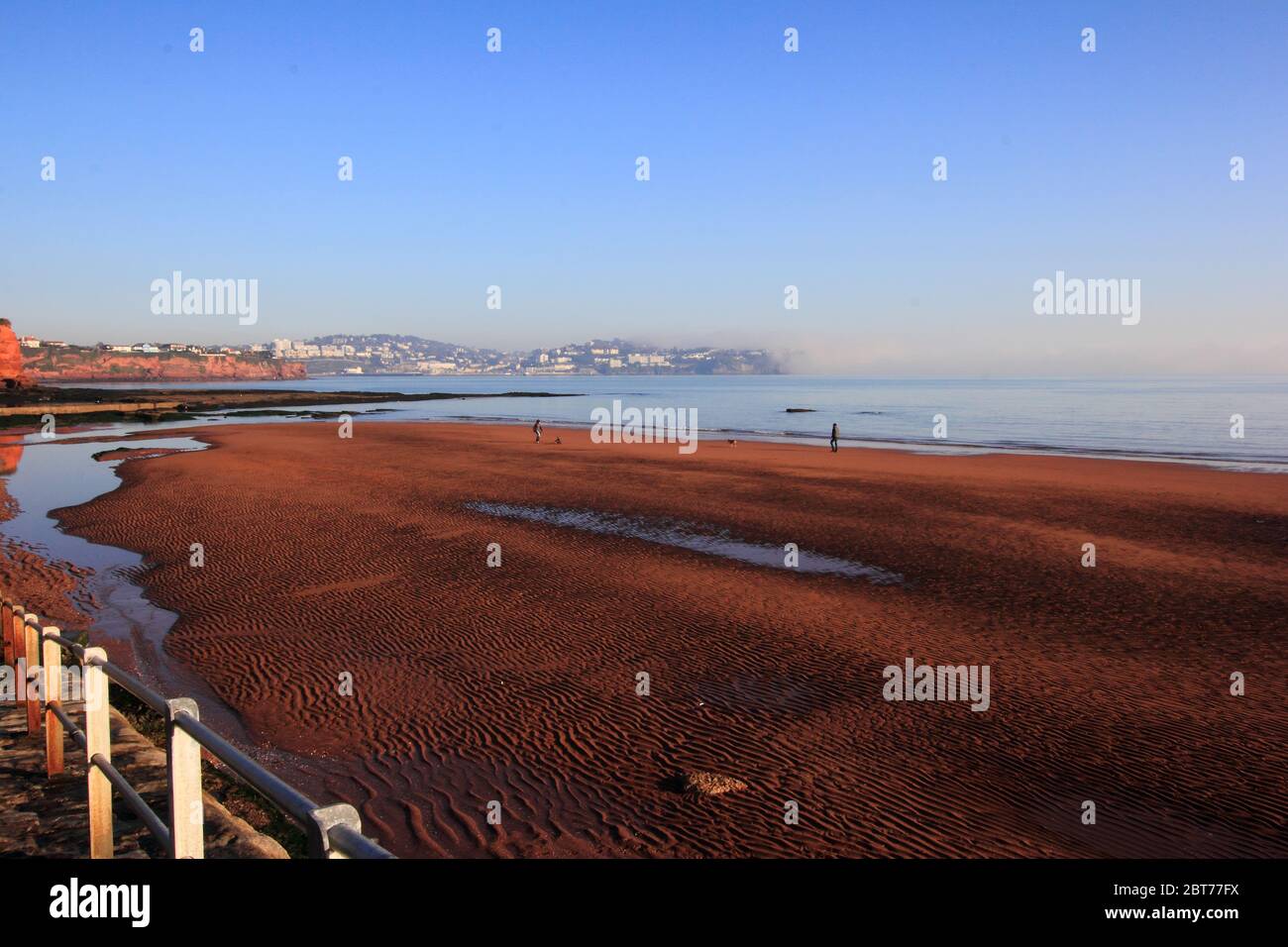 I Dog Walker sulla spiaggia di sabbia rossa increspata, Preston Sands, Paignton, Torbay con le scogliere di arenaria rossa che lavorano intorno a Torquay parzialmente avvolte in un mare Foto Stock