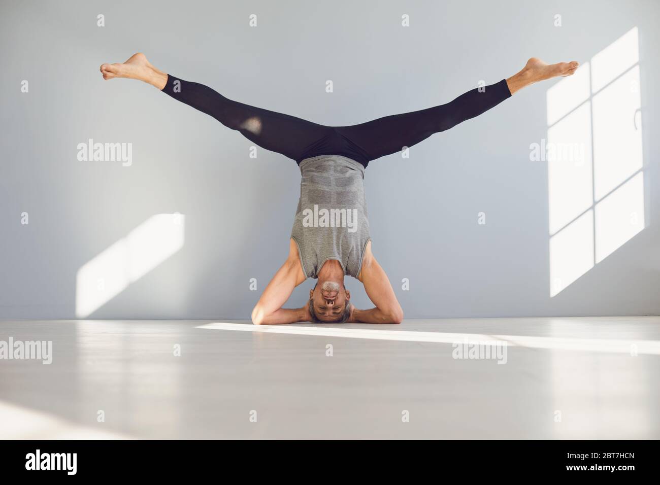 Adulto a piedi nudi maschio in abbigliamento sportivo che esegue l'esercizio di yoga sirsasana sul pavimento in studio luce contemporaneo contro parete bianca Foto Stock