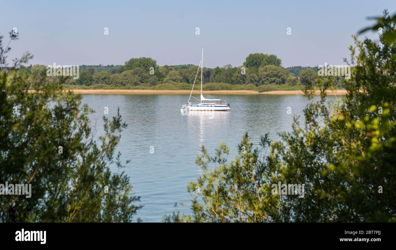 Paesaggio romantico con una barca a vela di colore bianco ancorata ad un lago (Ammersee). Foglie di albero in primo piano. Formato panorama. Foto Stock