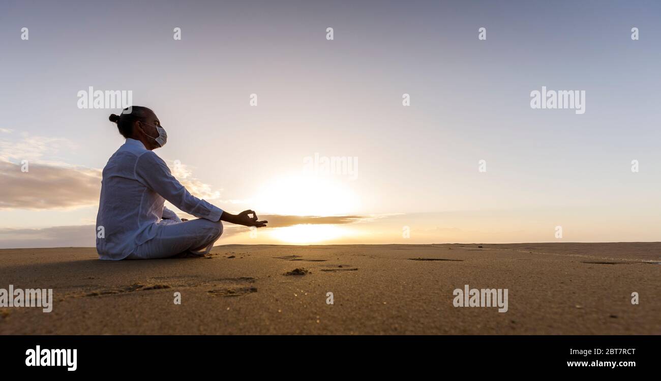 meditando l'uomo in maschera medica in lotus posa sulla spiaggia, maschio indossando maschera chirurgica viso e bianco yoga vestiti con top knot acconciatura all'alba Foto Stock