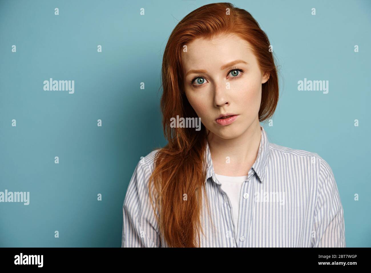 Colpo di testa. Una ragazza dai capelli rossi con occhi blu in una camicia a righe si erge su uno sfondo blu e guarda nella fotocamera. Foto Stock