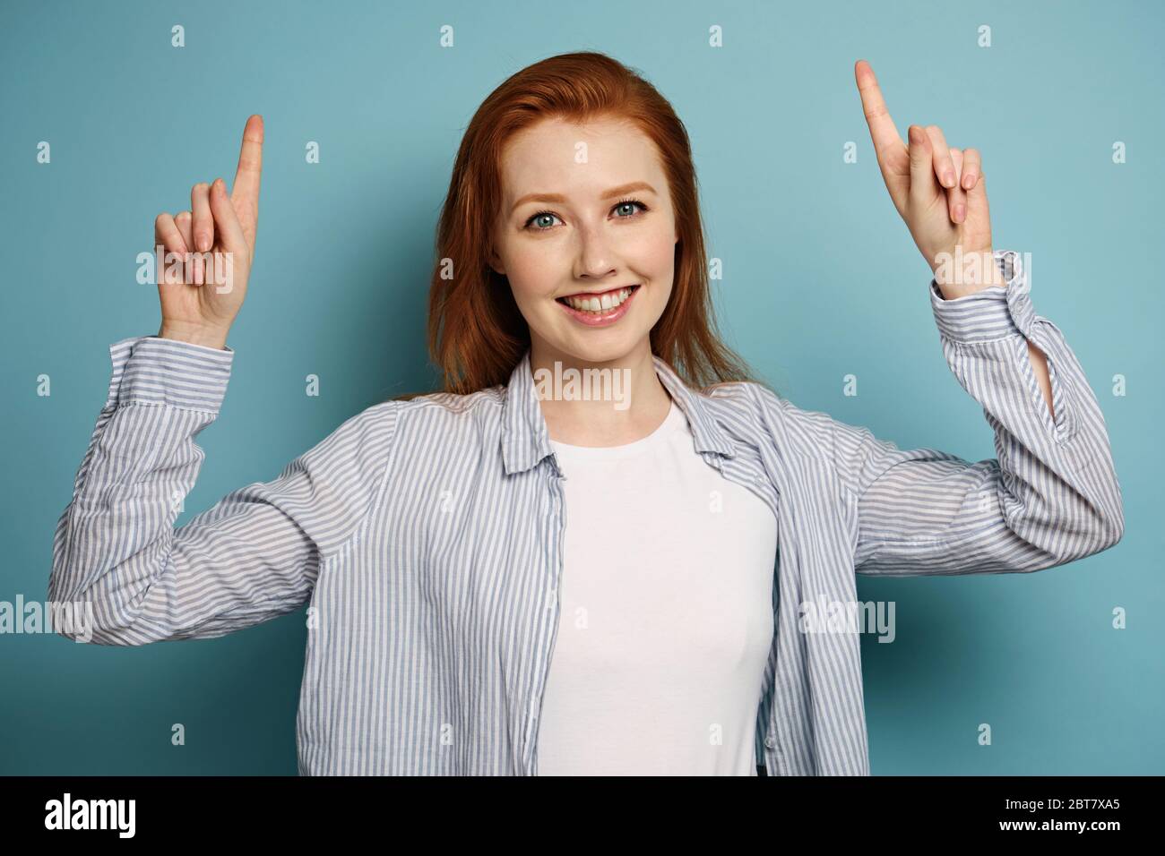 La ragazza dai capelli rossi in una camicia a righe sorride e punta le dita verso la parte superiore in piedi su uno sfondo blu. Foto Stock