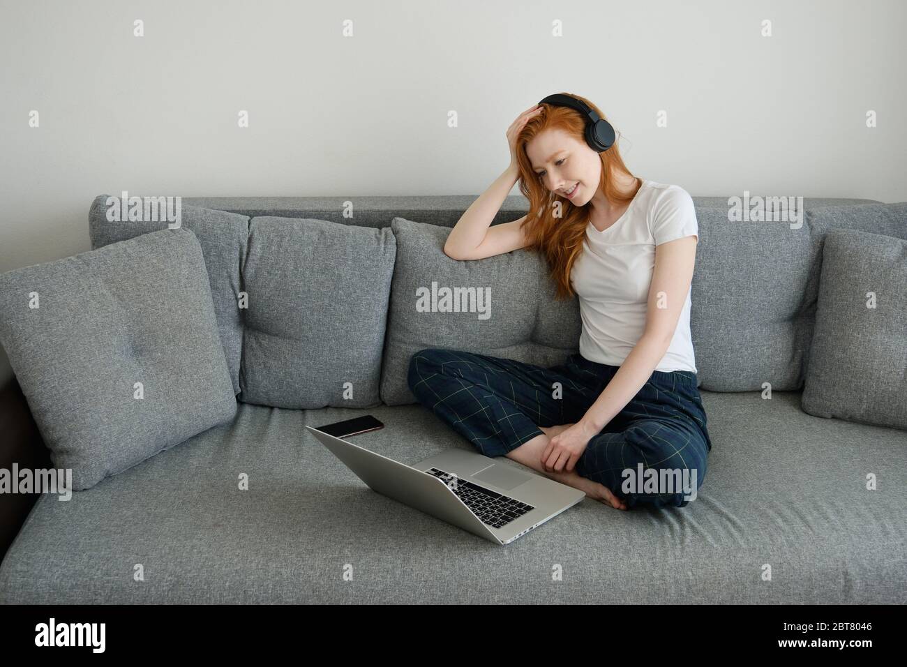 Ragazza rossa seduta sul divano con le gambe incrociate in abiti e cuffie e sorridente, guardando il lapto Foto Stock
