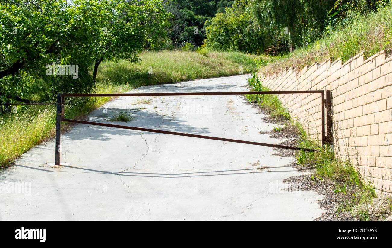 un cancello in metallo arrugginito che blocca una strada abbandonata che si dirige su una collina alberata con un muro di un blocco di scorie in una giornata di sole Foto Stock