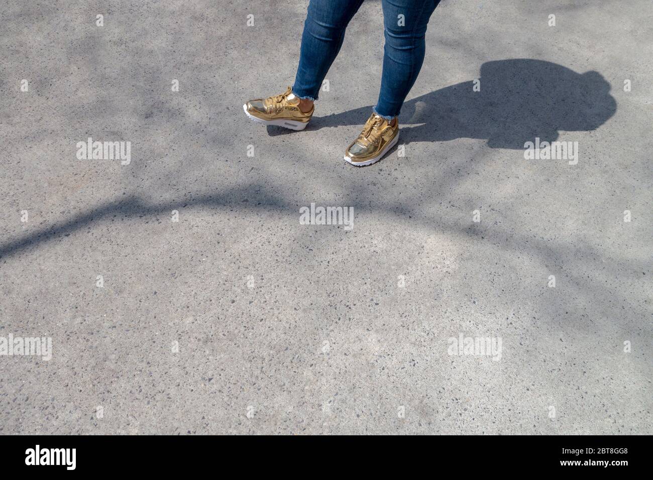 Donna leggermente sovrappeso con jeans blu skinny e sneakers di moda bianco dorato che aspettano in piedi su un passaggio in una bella giornata di sole con ombra e t Foto Stock