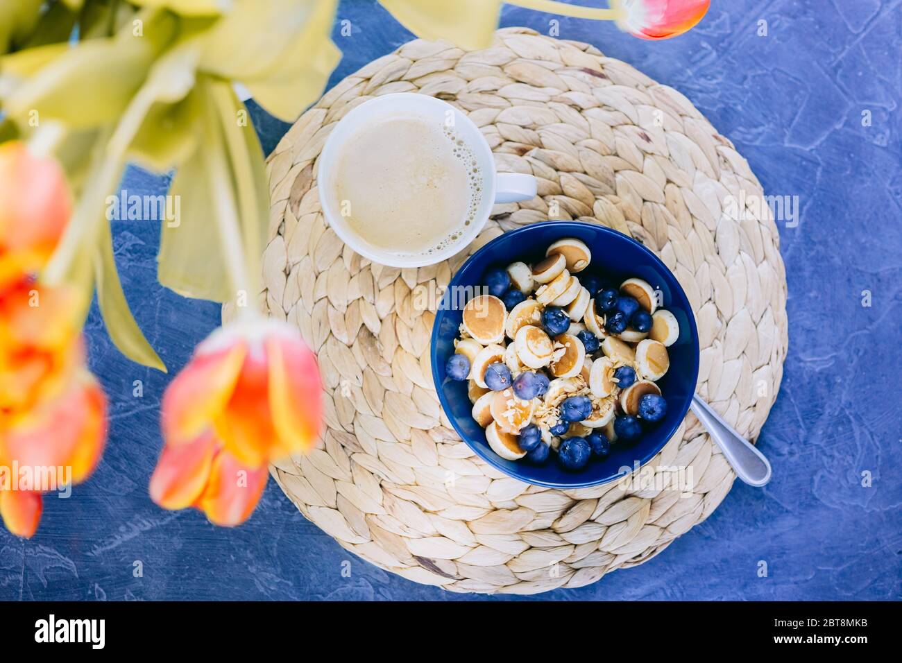 Mattina, colazione, cereali, mini pancake, mini pancake in una ciotola blu scuro con miele di sciroppo d'acero con mirtillo e una tazza di caffè. St Foto Stock