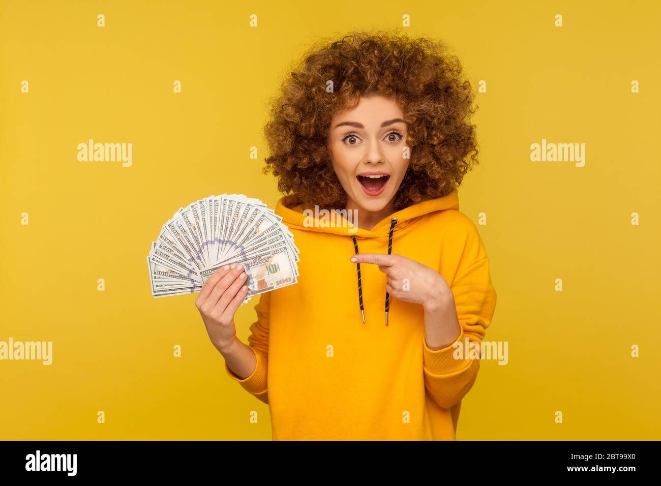 Ritratto di sorpresa gioiosa donna riccamente-capelli in felpa di stile urbano che punta a mazzo di banconote del dollaro, guardando scioccato dalla fortuna nella lotteria, vantare Foto Stock