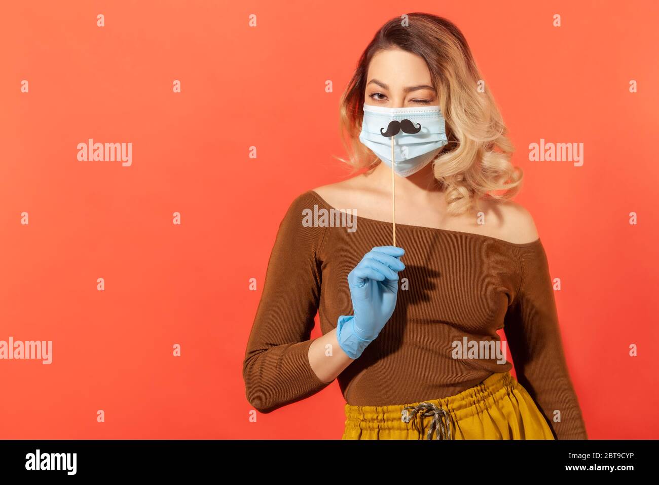 Ritratto di donna positiva in guanti protettivi, tenendo falso accessorio baffi sopra maschera chirurgica viso, winking giocosamente, di buon umore in nonostante il decor Foto Stock