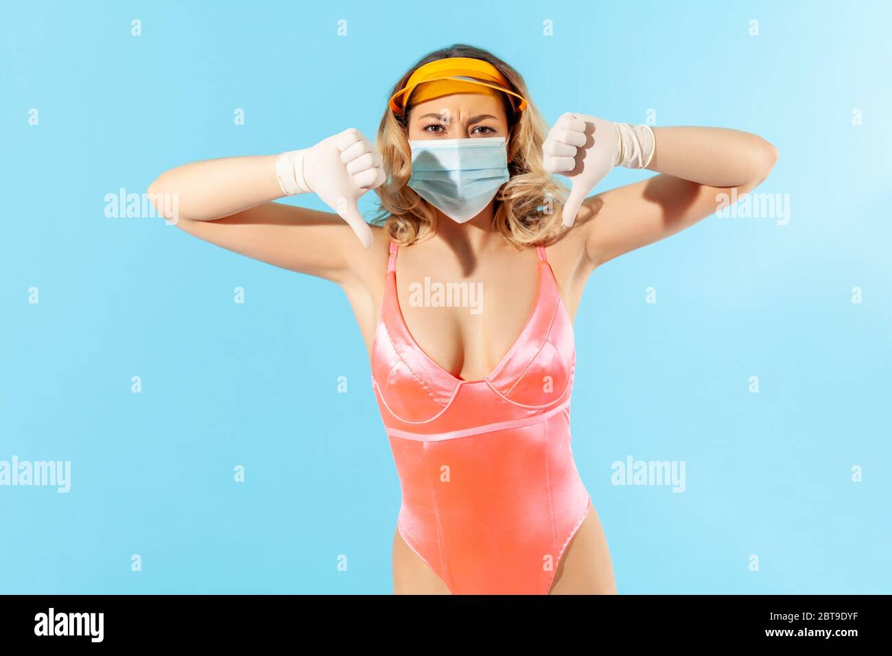Donna dispiaciuta in costume da bagno, indossando maschera igienica per il viso e guanti chirurgici per prevenire il coronavirus contagioso in resort, mostrando pollici verso il basso, non piace Foto Stock
