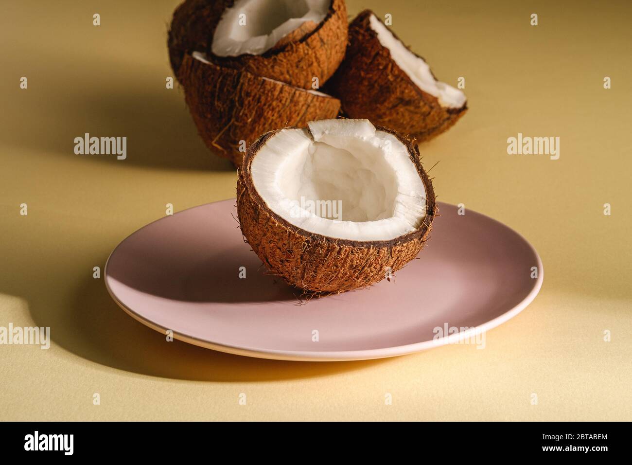 Noce di cocco mezzo in piatto rosa con frutta a guscio su crema giallo fondo pianura, astratto cibo tropicale concetto, angolo vista Foto Stock