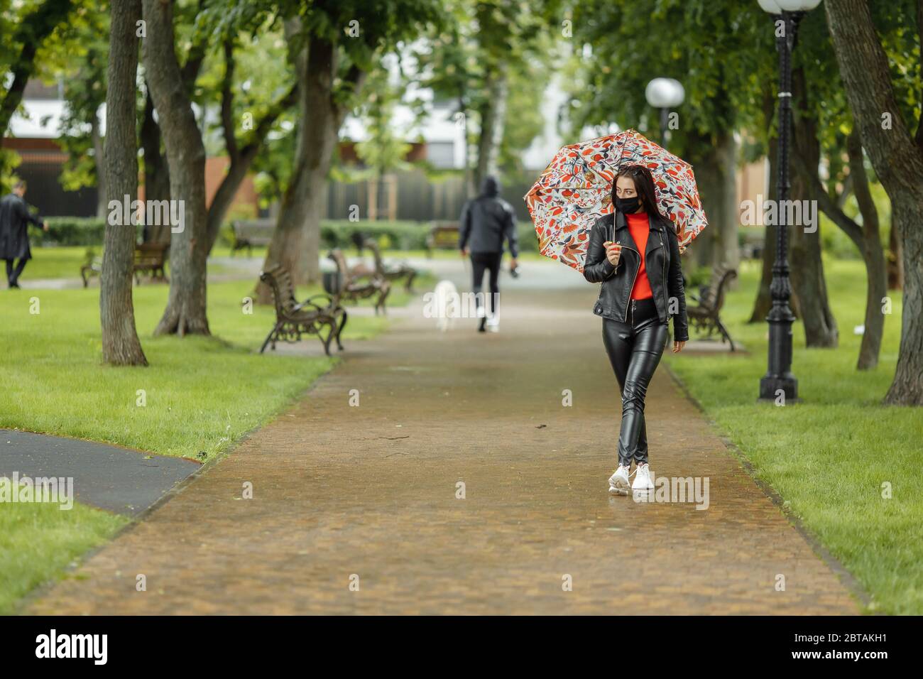 Una ragazza mascherata sta camminando lungo la strada. Una ragazza in una maschera protettiva cammina nel parco con un ombrello sotto la pioggia. Infezione da coronavirus COVID-19 Foto Stock