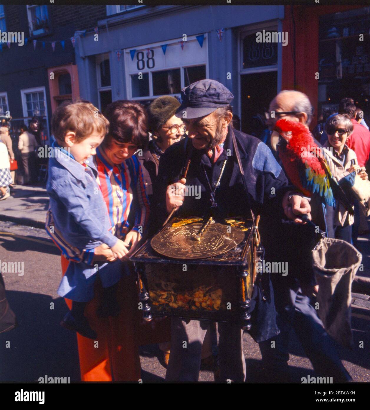 Un busker di strada con la sua scatola di musica e pappagallo intrattiene un ragazzo e la folla nel mercato antico di Portobello Road a Notting Hill, Londra negli anni '70. Foto Stock