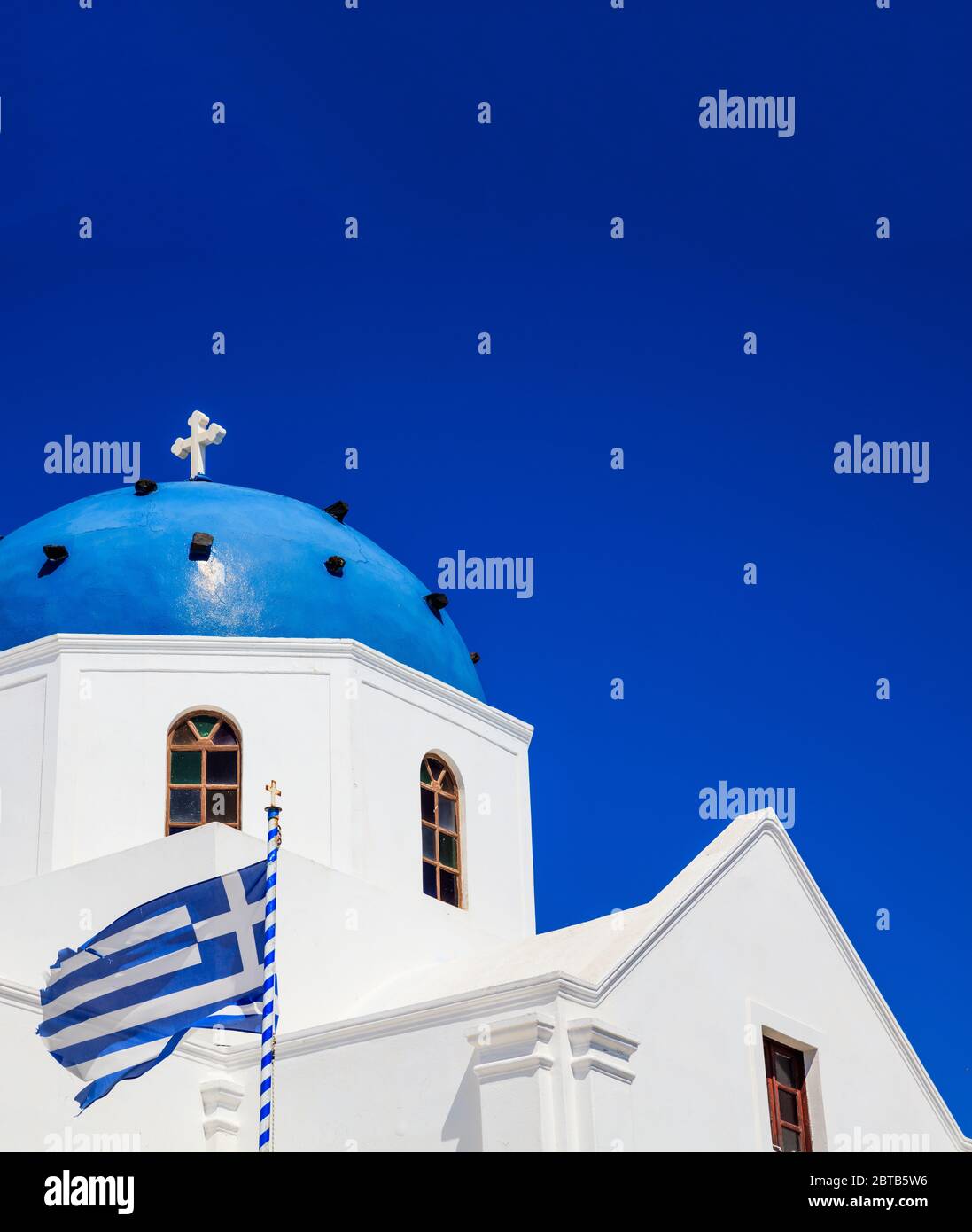 Isola di Santorini, Grecia. Bandiera greca che sventola sulla chiesa ortodossa bianca con cupola blu su sfondo blu cielo chiaro, foto verticale Foto Stock