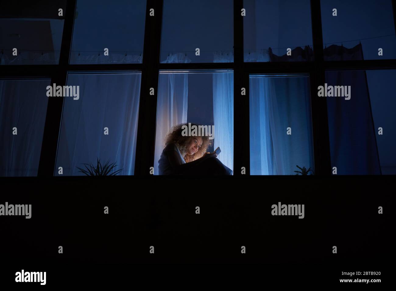 Vista laterale grandangolare su una giovane donna dai capelli ricci seduta sulla finestra una notte e utilizzando smartphone, spazio per la copia Foto Stock