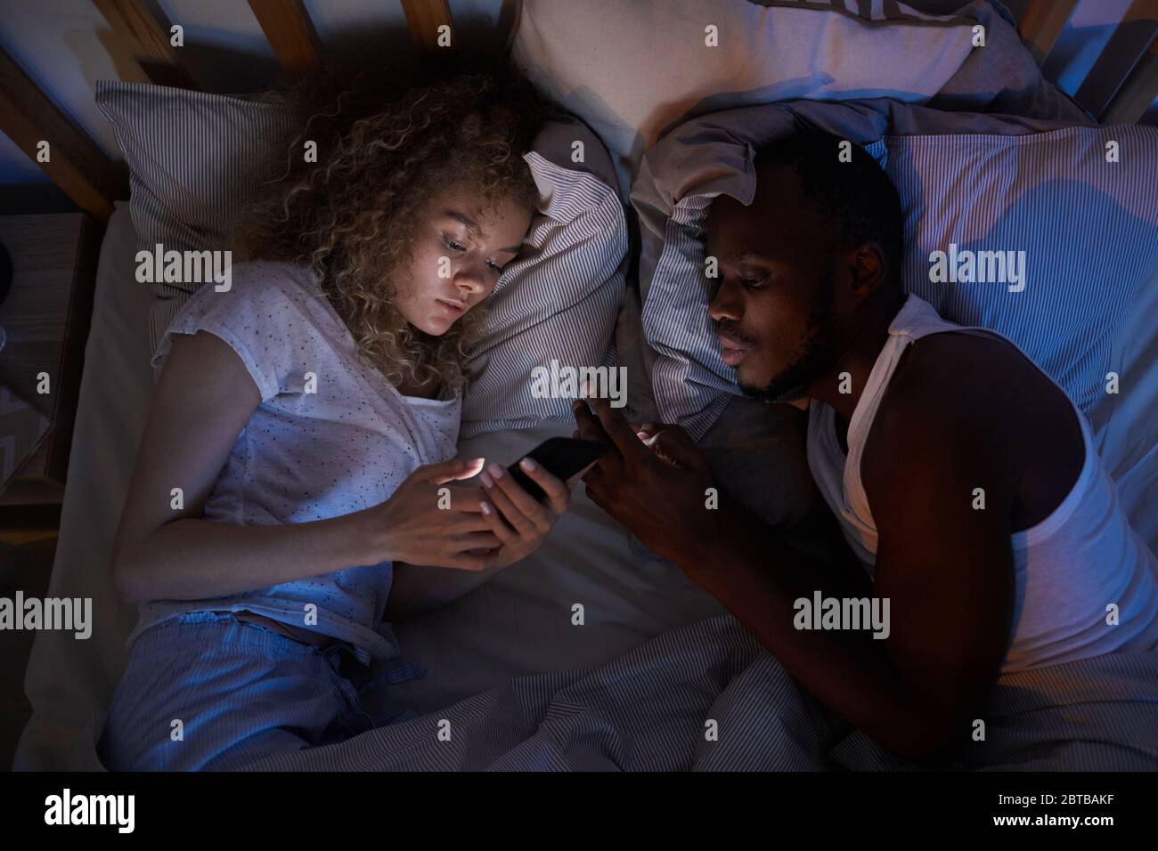 Vista sopra a giovane coppia mista-corsa sdraiata a letto di notte e utilizzando smartphone, copia spazio Foto Stock