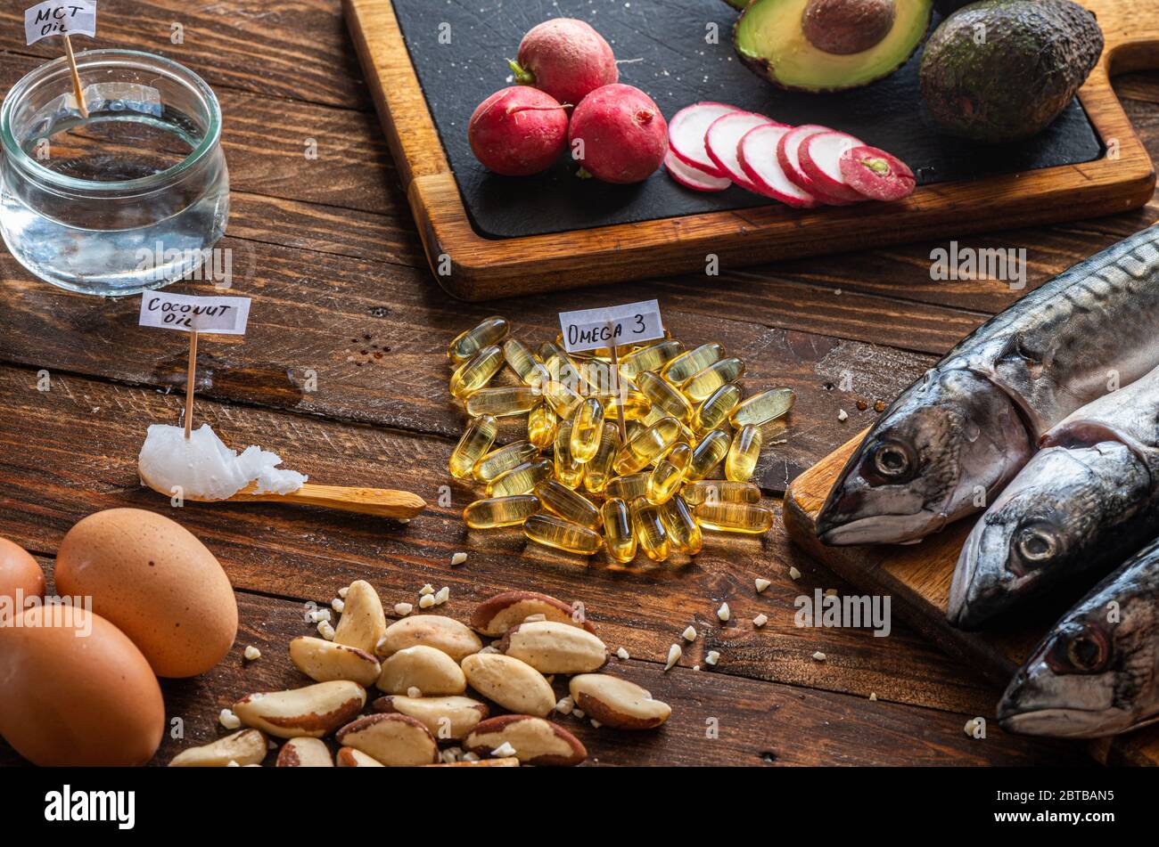 Primo piano di alimenti chetogenici: Olio MCT, pesce sgombro, avocado, uova, burro di cocco, noci brasiliane, ravanelli, supplementi Omega-3 su un tavolo di legno Foto Stock