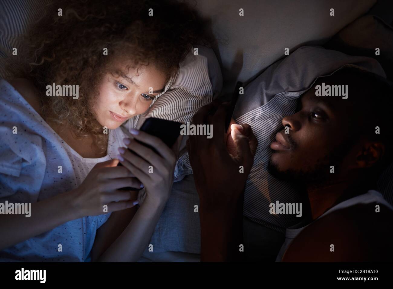 Sopra la vista di una giovane coppia mista che usa gli smartphone mentre si sdraia a letto di notte, concentrati sulla donna sorridente dai capelli ricci Foto Stock