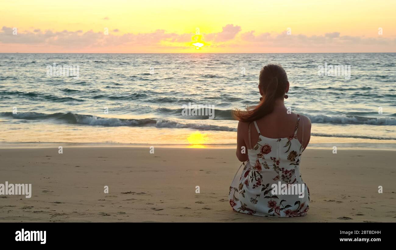 giovane donna in elegante abito estivo si siede sulla spiaggia di sabbia guardando l'oceano pittorico al tramonto vista sul retro Foto Stock