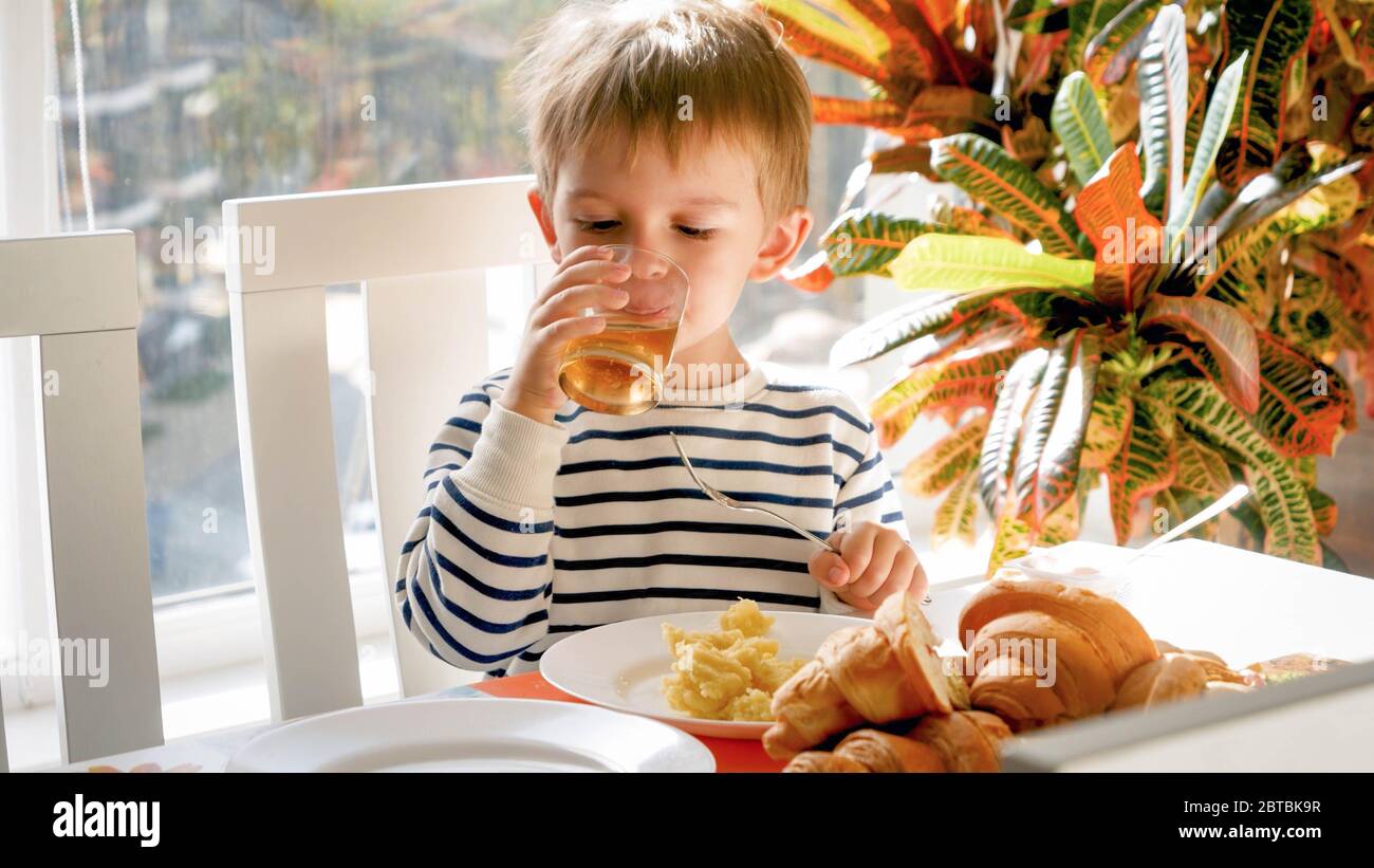 Bel ragazzo di 3 anni che beve succo di mela dal bicchiere durante la cena in sala da pranzo Foto Stock