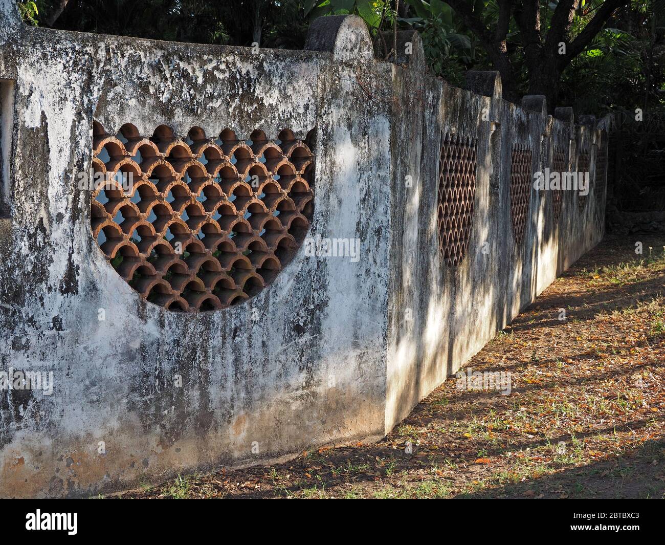 Antico muro in cemento dipinto di bianco con aperture in cotto nella luce della sera con alberi tropicali sullo sfondo a Malindi sulla costa del Kenya, Africa Foto Stock