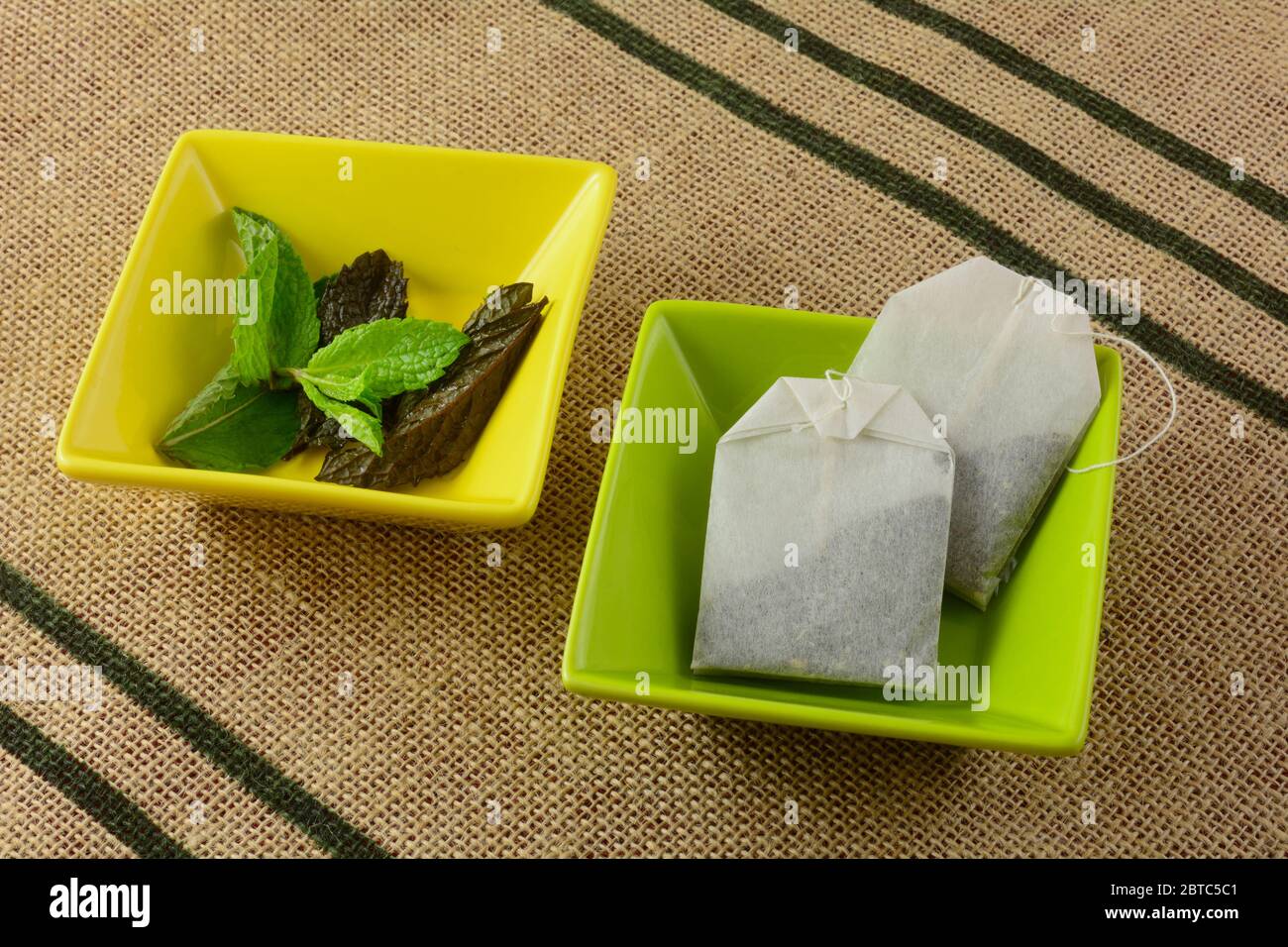 Due sacchetti di tè alla menta in una ciotola verde e foglie di menta fresca in una ciotola gialla sul tappetino Foto Stock