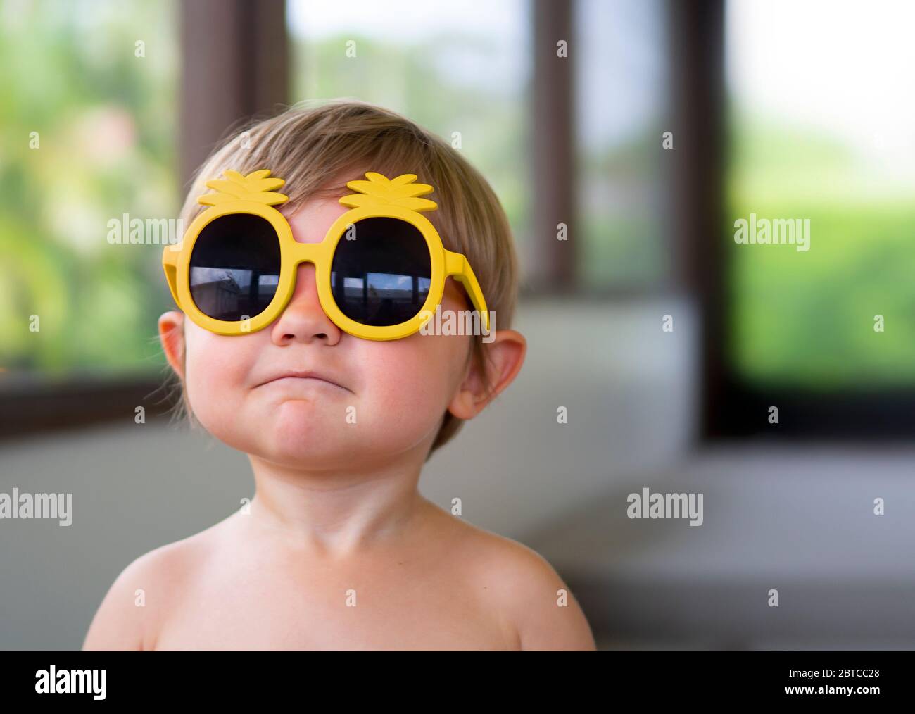 Ritratto di un ragazzo di due anni che indossa occhiali da sole gialli guardando nella macchina fotografica Foto Stock
