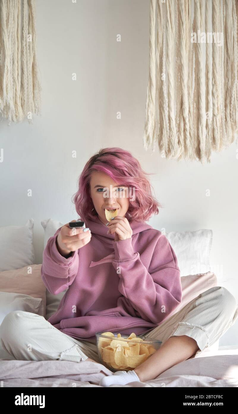 Felice Hipster gen z ragazza teen con capelli rosa guardando la tv mangiare chip a letto. Foto Stock