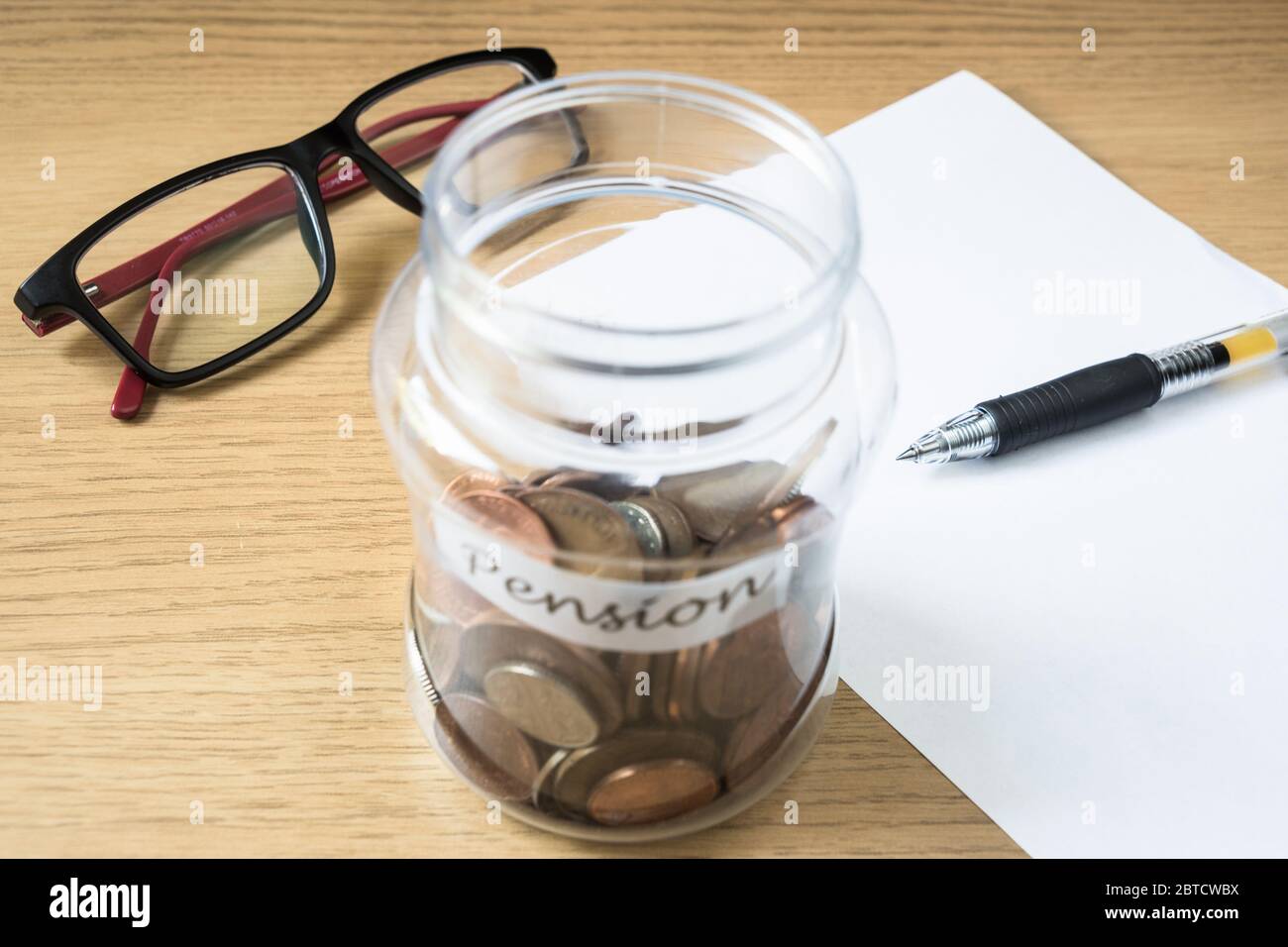 Risparmio di moneta in un vaso come banca piggy verso pensione con bicchieri penna e carta sul tavolo Foto Stock