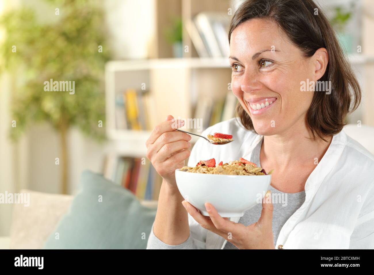 Donna adulta felice che tiene la ciotola dei cereali guardando via facendo colazione seduto nel divano nel soggiorno a casa Foto Stock