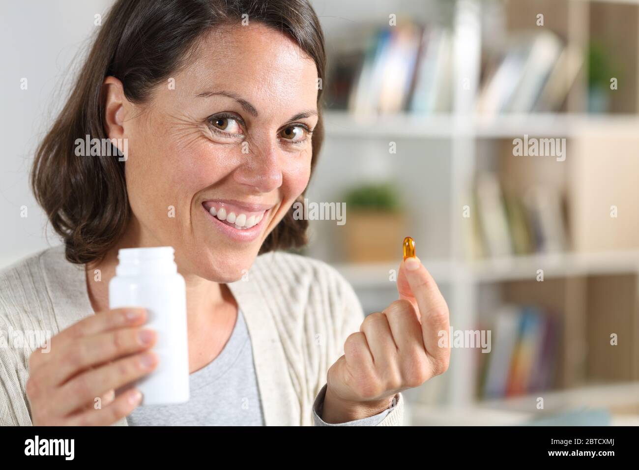 Donna adulta felice che mostra la pillola di vitamina che tiene il flacone guardando la macchina fotografica a casa Foto Stock