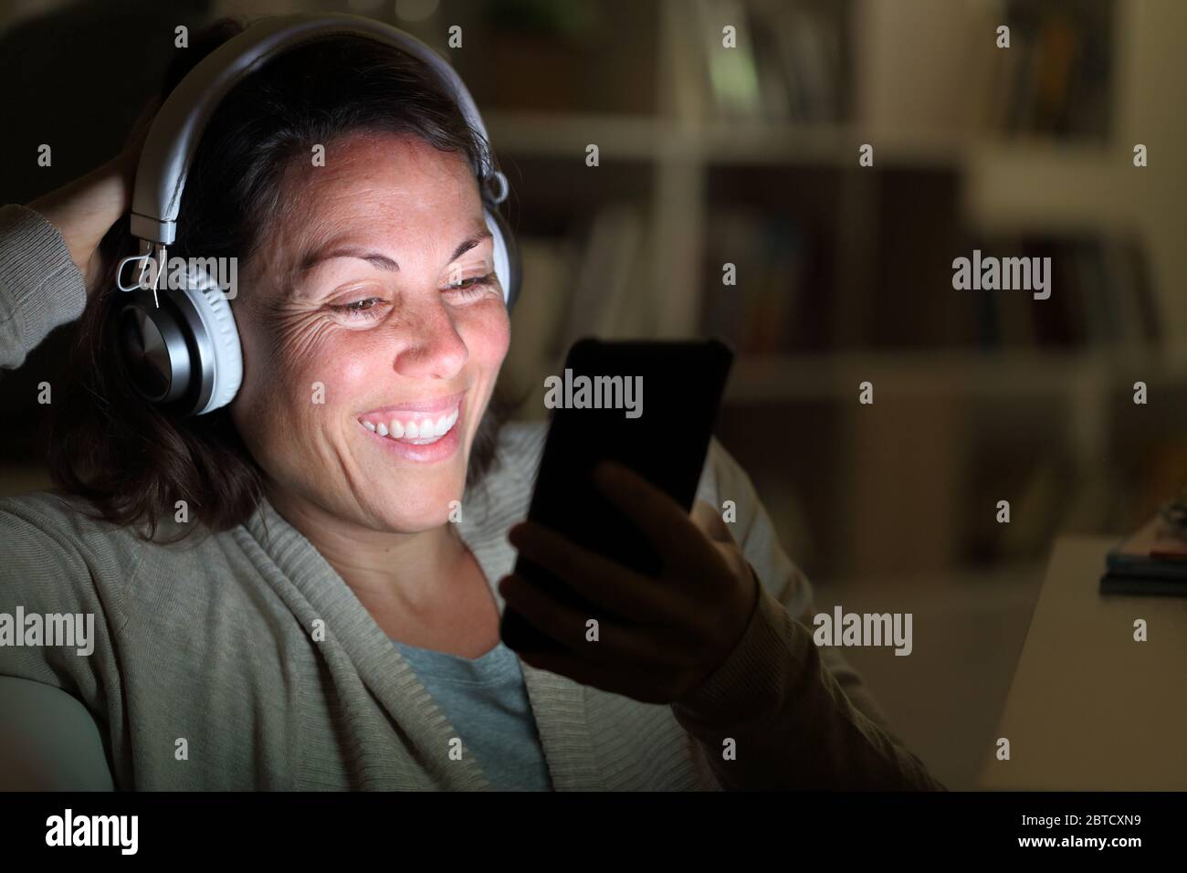 Donna adulta felice con le cuffie ascolta la musica sullo smartphone con schermo illuminato in salotto di notte a casa Foto Stock