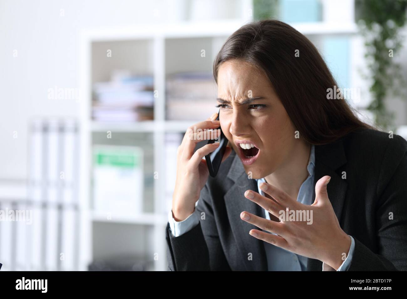 Donna dirigente arrabbiata che si è addetta a chiamare uno smartphone seduto su una scrivania in ufficio Foto Stock