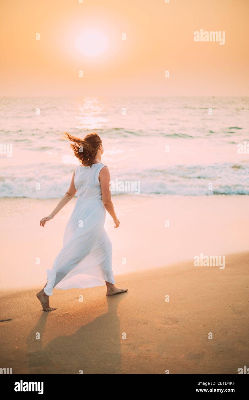 Goa, India. Giovane donna caucasica con capelli fluttering in vento in Bianco Abito camminare lungo Seashore in Alba tempo di tramonto. Foto Stock