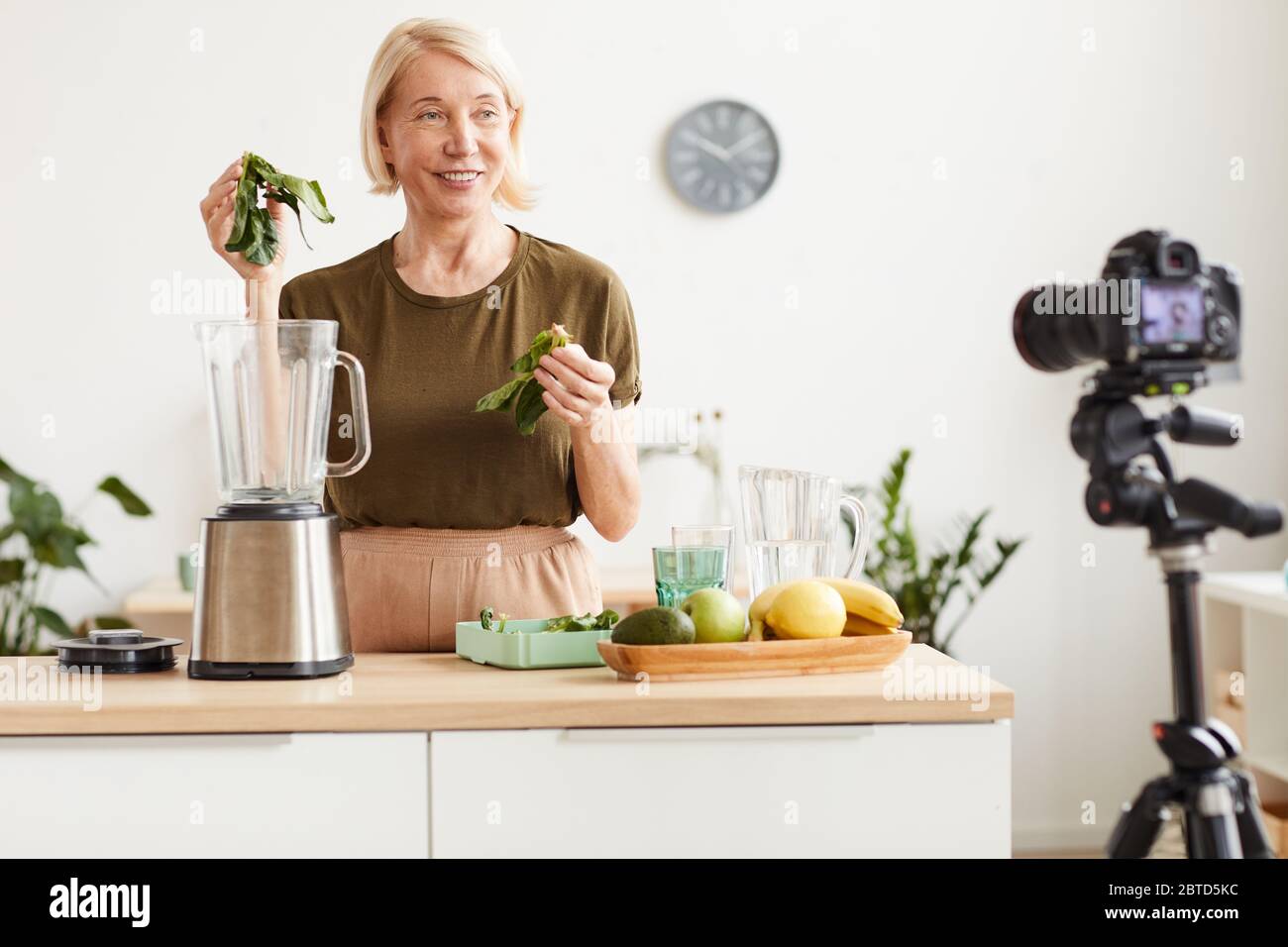Il blogger del cibo sorridente alla macchina fotografica e mostrando il processo di fare un cocktail sano in cucina Foto Stock