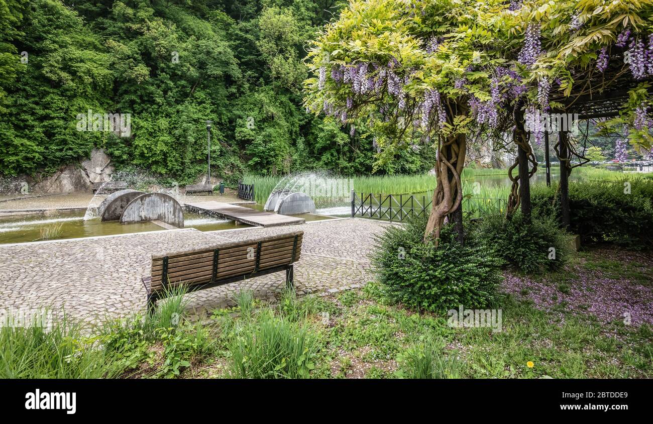 Panca vuota nel parco con una pianta di glicine e una fontana moderna vicino al centro della città di Durbuy nelle Ardenne belghe. Foto Stock