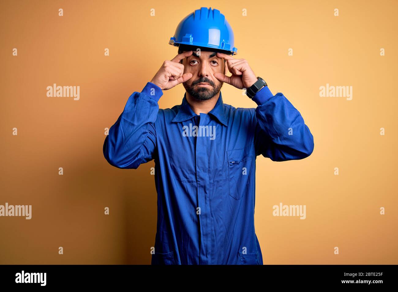 Uomo meccanico con barba che indossa uniforme blu e casco di sicurezza su sfondo giallo cercando di aprire gli occhi con le dita, sonnolento e stanco per mattina f Foto Stock