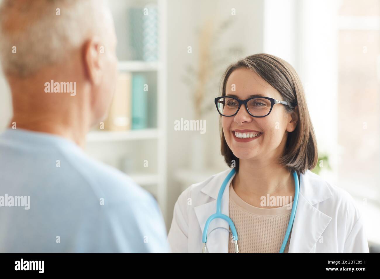 Ritratto di testa e spalle di donna sorridente che agita le mani con la paziente anziana mentre si trova in piedi all'interno della clinica Foto Stock