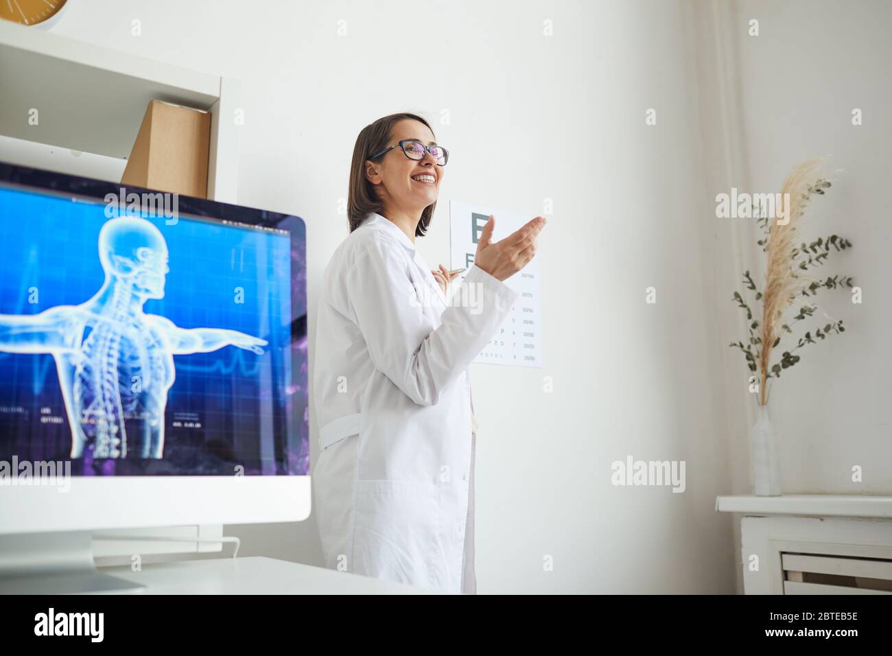 Vista laterale ritratto di un'oftalmologista sorridente che punta alla mappa durante il test oculare in clinica, spazio di copia Foto Stock